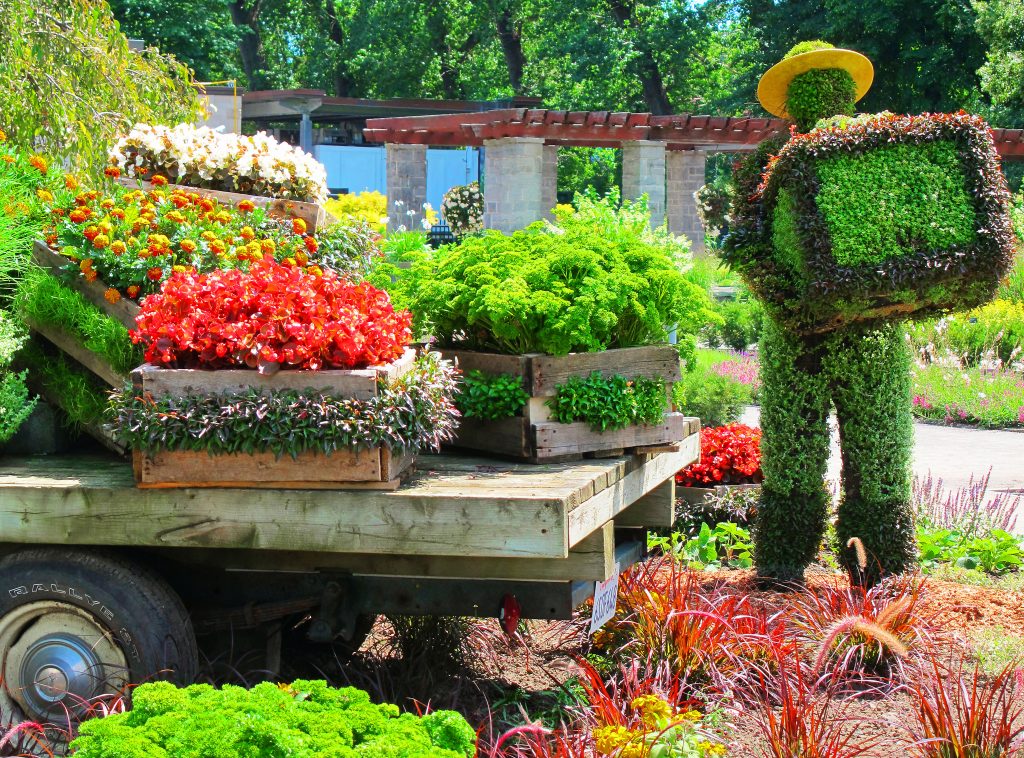 A whimsical topiary figure donning a hat seems to be tending to a cart overflowing with colorful flowers and plants in a lush garden gallery setting.