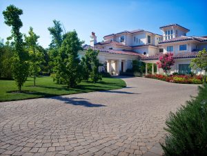 A grand, luxurious home featuring a cobblestone driveway, surrounded by meticulously landscaped greenery and bright pink bougainvillea under a clear blue sky offers inspiration on how to create a beautiful landscape