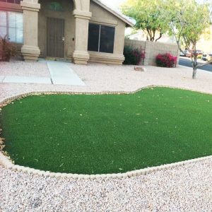 A neatly maintained front yard showcasing creative and unique landscape designs, with a section of artificial green grass surrounded by decorative rocks, complementing a suburban home with a stucco exterior.