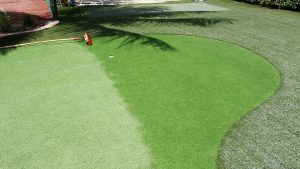 A mini-golf green with a putter and golf ball near the hole, surrounded by artificial turf and a playful shadow of a palm tree on a sunny day, is part of our Creative Landscape Designs
