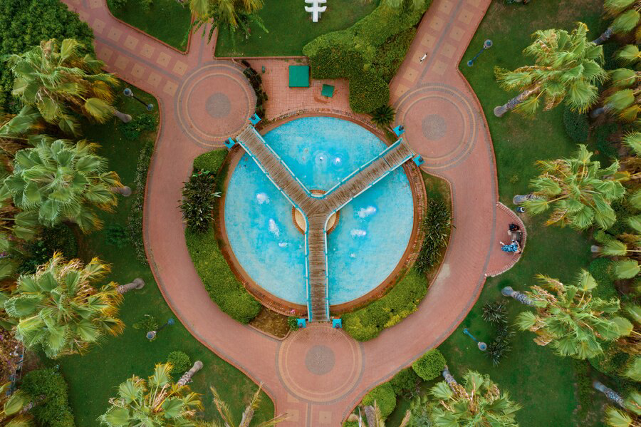 An aerial view of a symmetrical turquoise swimming pool, maximizing space and functionality with walkways resembling a pinwheel, surrounded by lush tropical foliage and brick pathways in a serene resort setting.