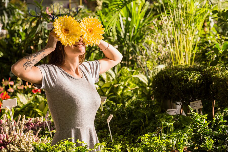 A joyful woman holding two bright yellow flowers over her eyes like glasses, surrounded by lush green plants in a seasonal splendor of a sunny garden atmosphere.