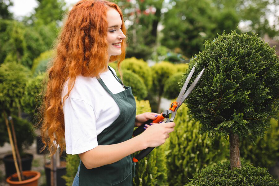 A smiling woman with red hair trims a bush with gardening shears, wearing a green apron and surrounded by lush plants.