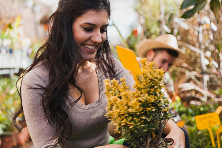 A smiling woman enjoying gardening as part of an eco-friendly landscape design, holding a potted plant with lush yellow blooms, with a person wearing a straw hat visible in the background, possibly assisting her.