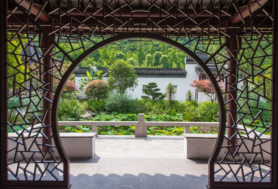 A tranquil Chinese garden viewed through an ornate, circular lattice window, inviting peace, contemplation, and embracing diversity.
