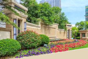 A colorful urban garden embodying Landscape Design Trends for 2024, with a variety of flowers blooming in the foreground, flanked by sandstone-clad walls and a small fountain, with modern