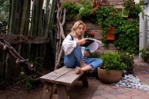 A woman engrossed in reading a book on "The Psychology of Landscape Design" while sitting cross-legged on a wooden bench in a tranquil garden oasis.