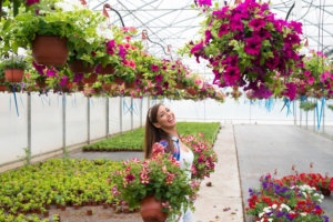 A joyful woman admiring vibrant hanging flowers in a sunlit greenhouse full of colorful blossoms, surrounded by the seasonal splendor.