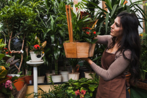 A woman with a brown apron admires a hanging basket of flowers in a lush greenhouse nursery, showcasing the concept of maximizing small spaces.