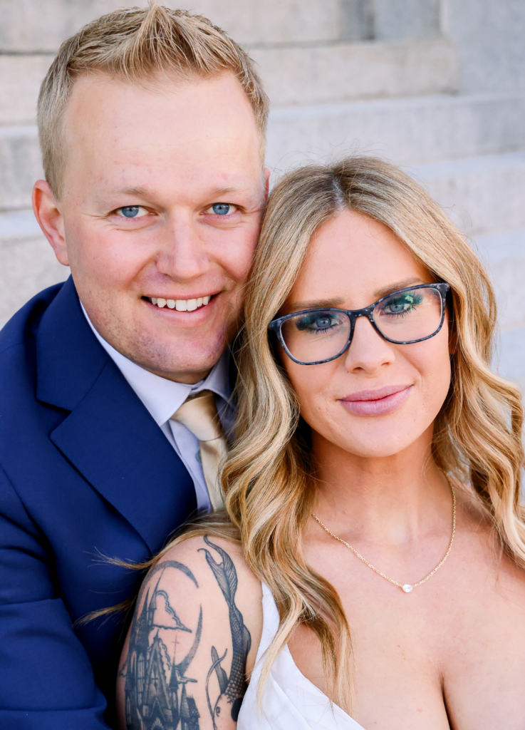 A man in a navy blue suit and a woman with wavy blonde hair and glasses, wearing a white dress, pose closely together in front of stone steps. The woman, showcasing her ship tattoo on her upper arm and donning a gold necklace with a small heart pendant, embodies the essence of our "About Us" story.