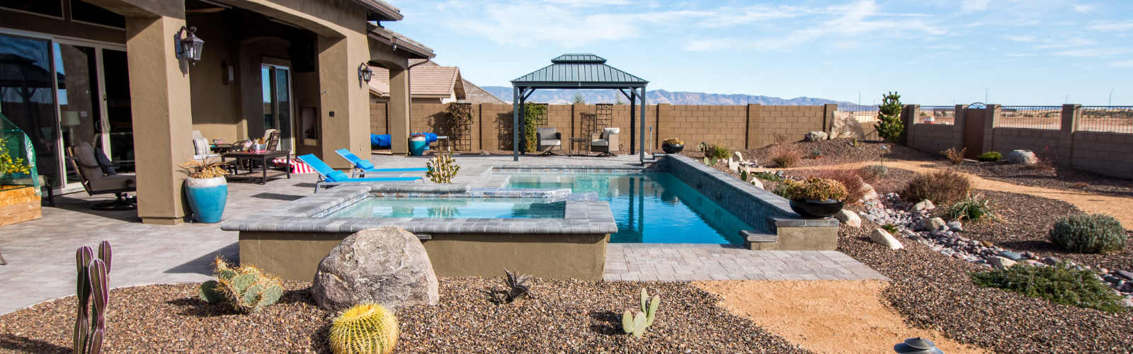 A modern backyard features a rectangular pool with an attached hot tub, surrounded by desert landscaping. There's a patio area with lounge chairs and a covered gazebo. The yard is enclosed by a brick wall, with mountains visible in the distance under a clear sky.