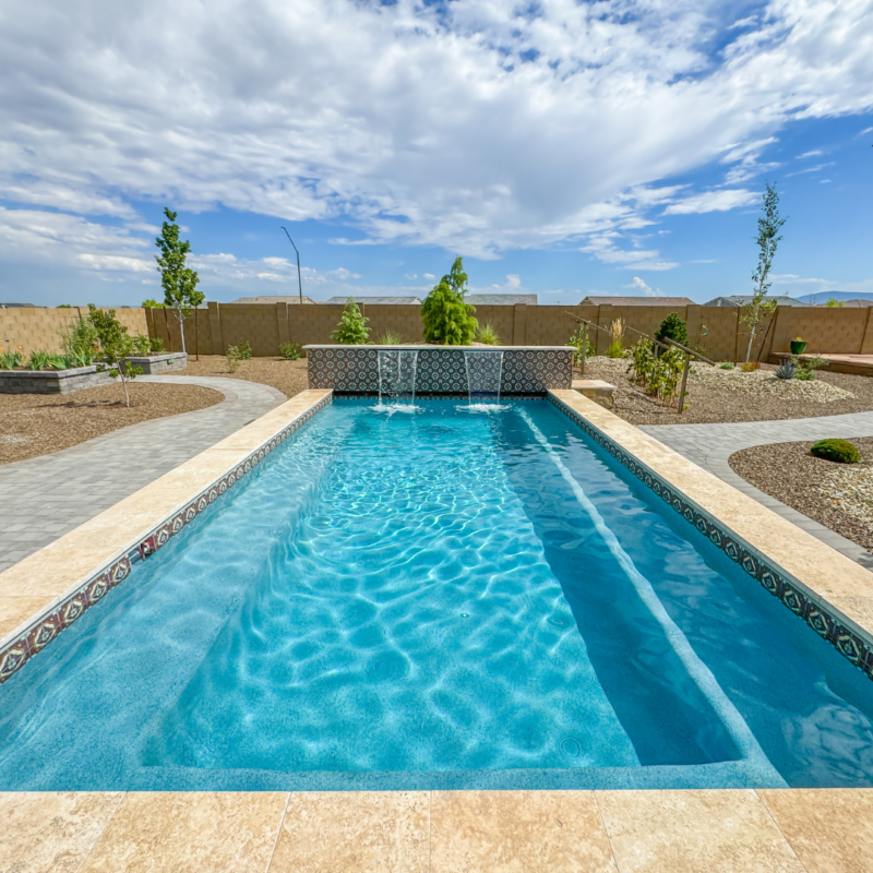 A rectangular swimming pool with clear blue water is featured in the backyard of the Hutchinson residence, surrounded by a stone deck. The pool has a patterned tile border and two small fountains at one end. Enclosed by a fence, the backyard boasts a pathway and landscaped garden area under a partly cloudy sky.