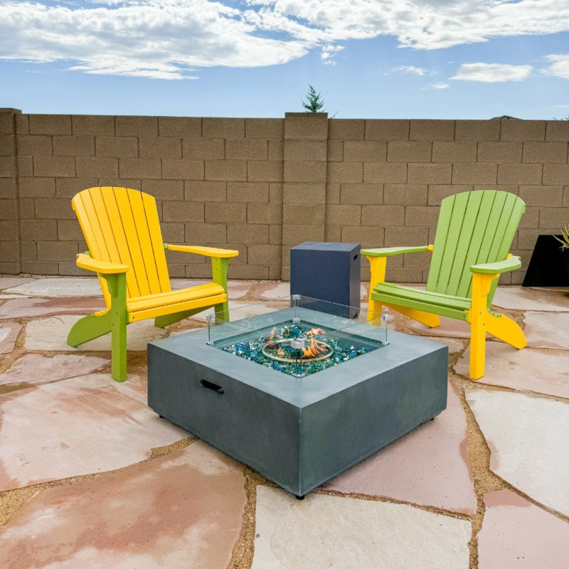 Two brightly colored Adirondack chairs, one yellow and one green, sit on a stone patio facing a modern square fire pit with glass pebbles. The setup at the Hutchinson Residence is enclosed by a tall beige brick wall under a blue sky with scattered clouds.