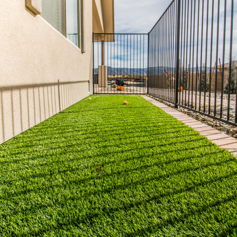 A small enclosed outdoor area with artificial grass and a metal fence on the right side sits beside the beige Hodges Residence. The area contains a single orange ball on the grass. In the background, construction materials are visible with mountains and cloudy skies beyond.