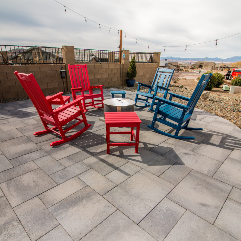 The Hodges Residence outdoor patio features stone pavers and four wooden rocking chairs around a small table. Two chairs and the table are red, while the other two chairs are blue. String lights hang above, with blurred houses and mountains in the background under a cloudy sky.