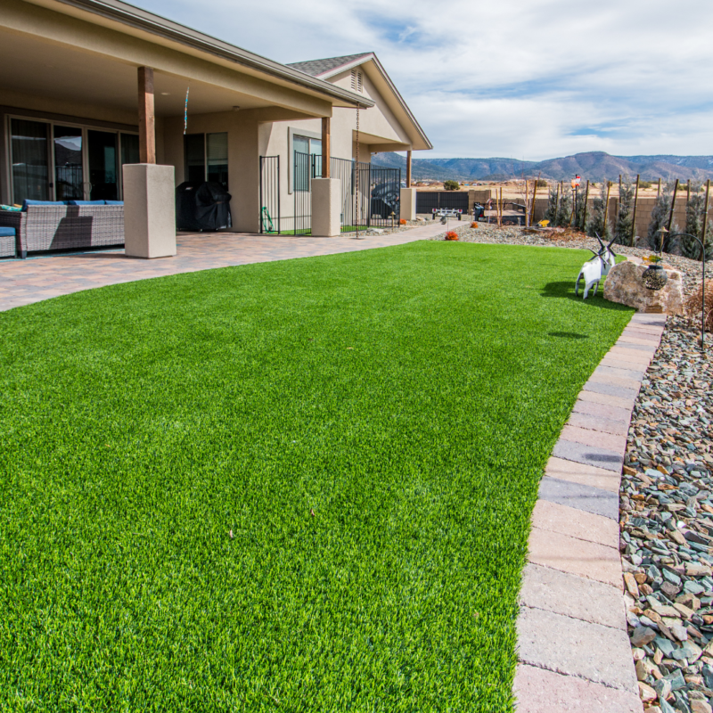 A modern residence with large windows and a covered patio is situated on a plot with a well-maintained green lawn. The lawn, bordered by a stone pathway and a rock garden, adds to the charm. Mountains are visible in the distance under a partly cloudy sky, completing this picturesque Hodges property.