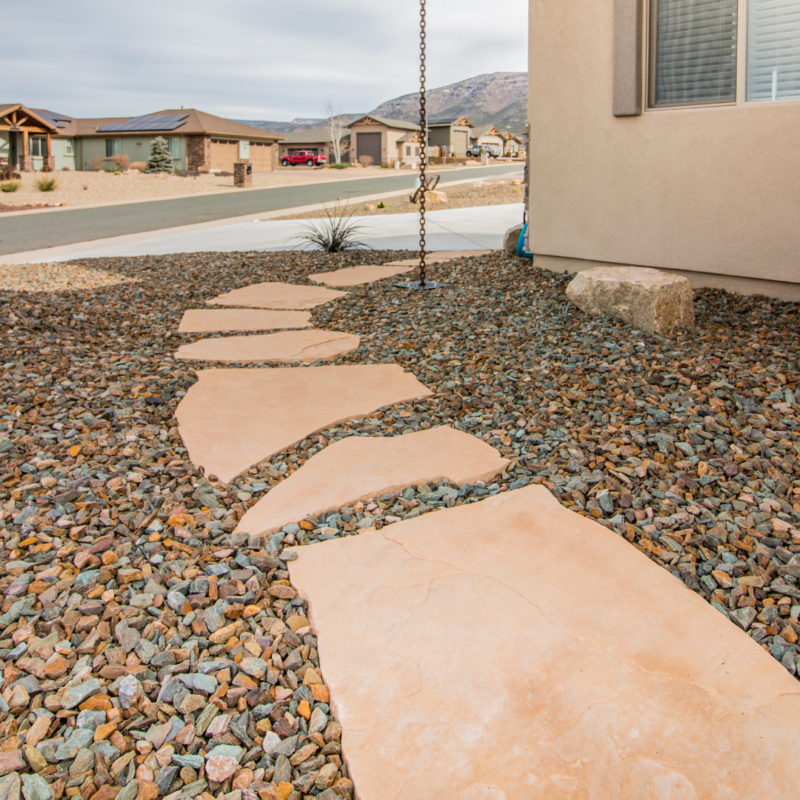 A pathway made of large, irregularly shaped stone slabs set in a landscape of small, multicolored rocks leads to the Hodges Residence in a desert-like neighborhood. The scene features a clear sky, distant mountains, and modern homes in the background.