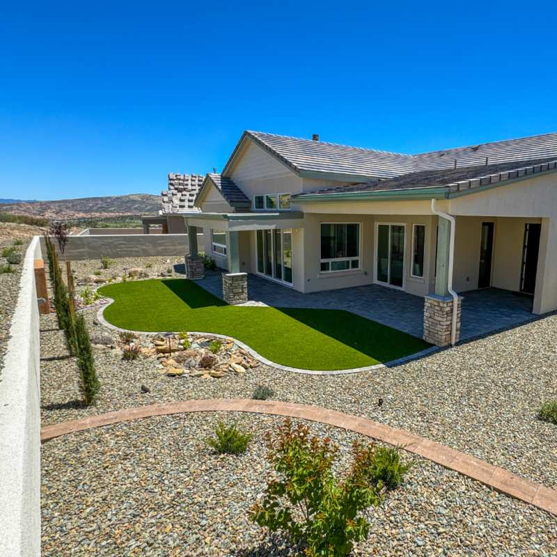 The Hampton Residence is a modern single-story home with a beige exterior, large windows, and a grey tile roof under a clear blue sky. The backyard features a neatly trimmed artificial lawn, a curved stone pathway, and a stone wall with scenic views of distant hills in the background.