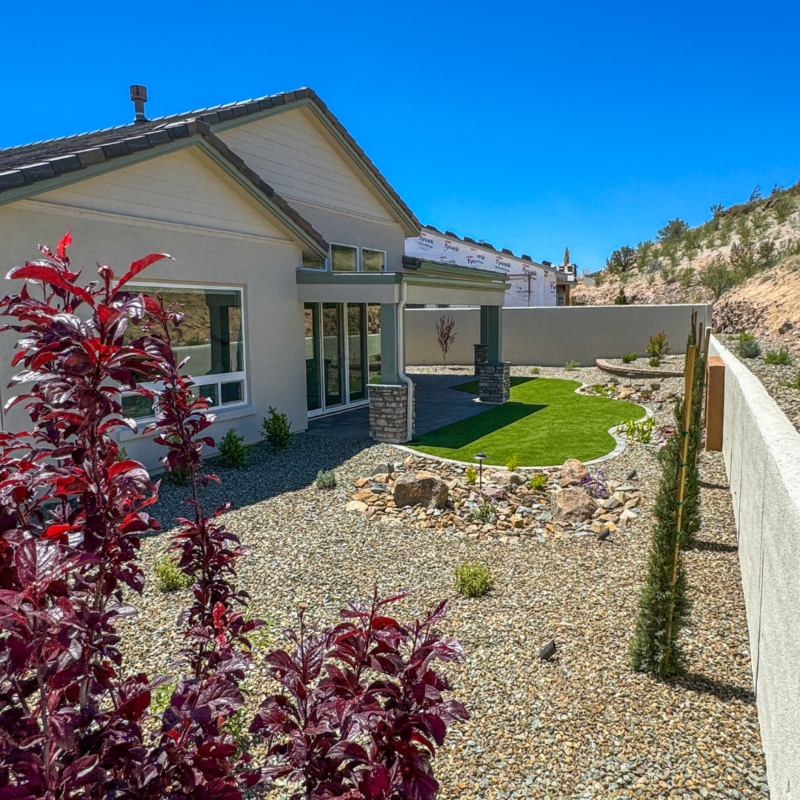 A Hampton residence with a light gray exterior sits under a clear blue sky. The backyard features a well-manicured lawn, gravel landscaping with a stone pathway, small shrubs, and a newly planted tree. A section of an adjacent unfinished property is also visible.