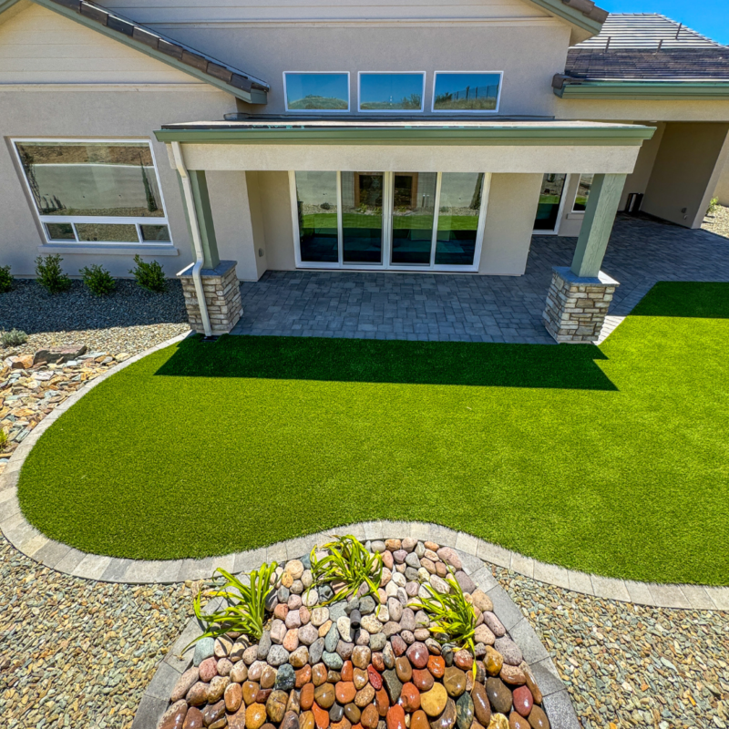 A modern Hampton residence with a well-maintained backyard features a covered patio with large windows and potted plants. The yard boasts vibrant green grass, surrounded by a rock landscaping walkway, and a circular area adorned with decorative stones.