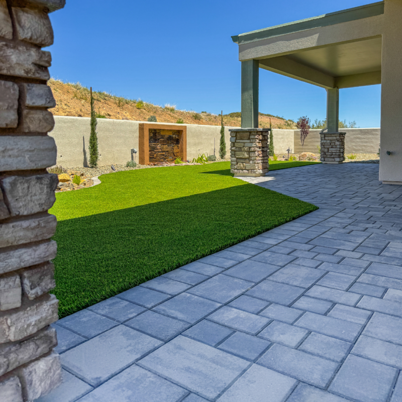 The Hampton Residence features a patio with gray pavers adjacent to a green artificial lawn, bordered by a stone wall with a built-in planter. The patio is partially shaded by a structure with stone pillars. In the background, the hill showcases sparse vegetation under a clear blue sky.