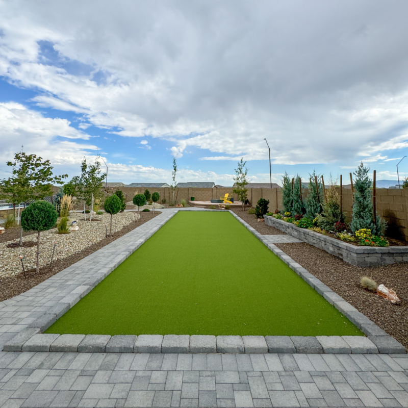 A well-maintained backyard at the Hutchinson residence features a long rectangular artificial grass area bordered by paver pathways and landscaped with various small shrubs, trees, and rocks. Two yellow chairs are positioned at the far end of the grassy area, and the sky is partly cloudy.