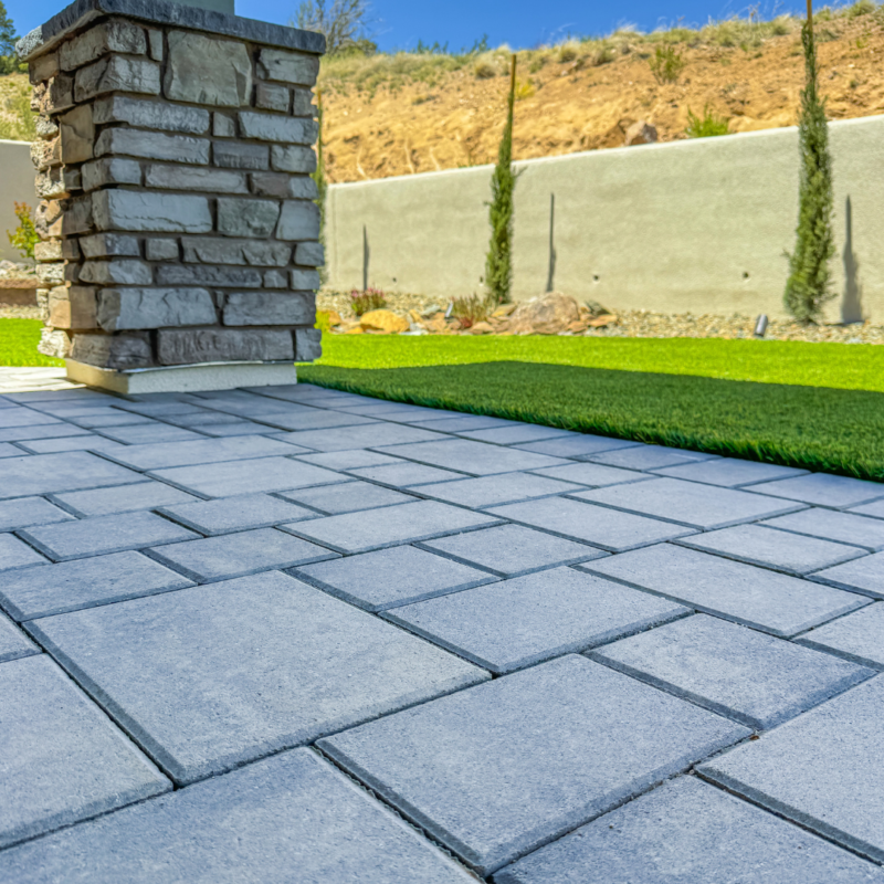 A modern backyard at the Hampton Residence features a stone-paved patio, a column covered in stacked stones, and a well-manicured lawn. The background showcases a light-colored wall, two tall, slender trees, and a rocky, shrub-filled landscape under a clear blue sky.