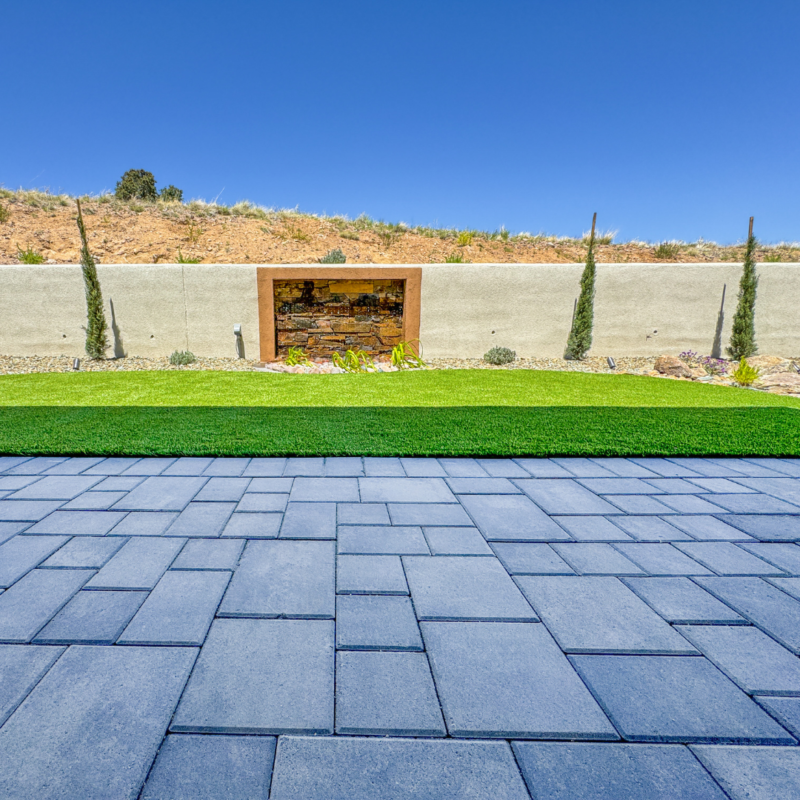 At the Hampton Residence, a modern backyard showcases a stone patio with large, flat pavers. Beyond the patio, there is a well-maintained lawn and a retaining wall with a built-in waterfall feature. The backdrop is a clear blue sky with some vegetation on a slight hill.