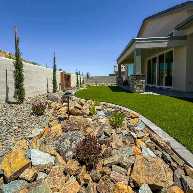 A modern Hampton backyard residence features a mix of artificial grass and a rock garden with various colored stones and small plants. Young trees line a white wall, and a patio area extends from the contemporary house with large windows and a blend of stone and glass.