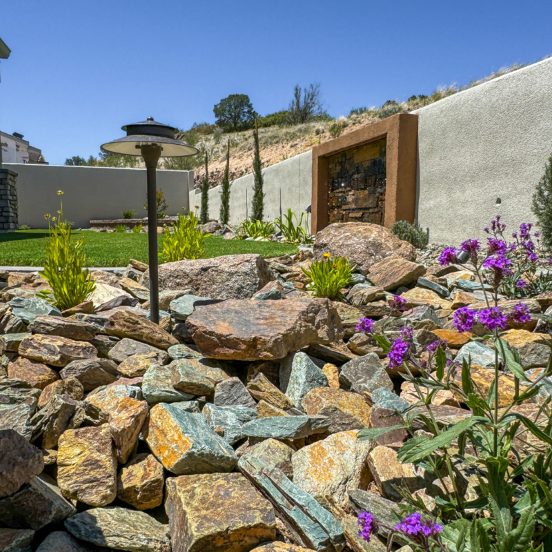 A serene garden scene at a Hampton residence with a rocky landscape featuring colorful stones, purple flowers, and green plants. A small, modern pathway light is visible on the left, and a square art piece is mounted on a white wall in the background under a clear blue sky.