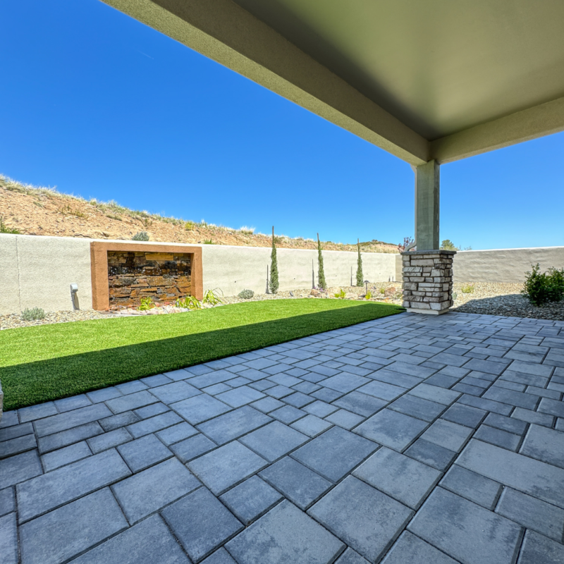 A covered patio with a paved floor overlooks a well-manicured lawn bordered by a stone wall, characteristic of a Hampton residence. The landscape includes several tall, slender trees and a small stone planter. The sky is clear and blue.