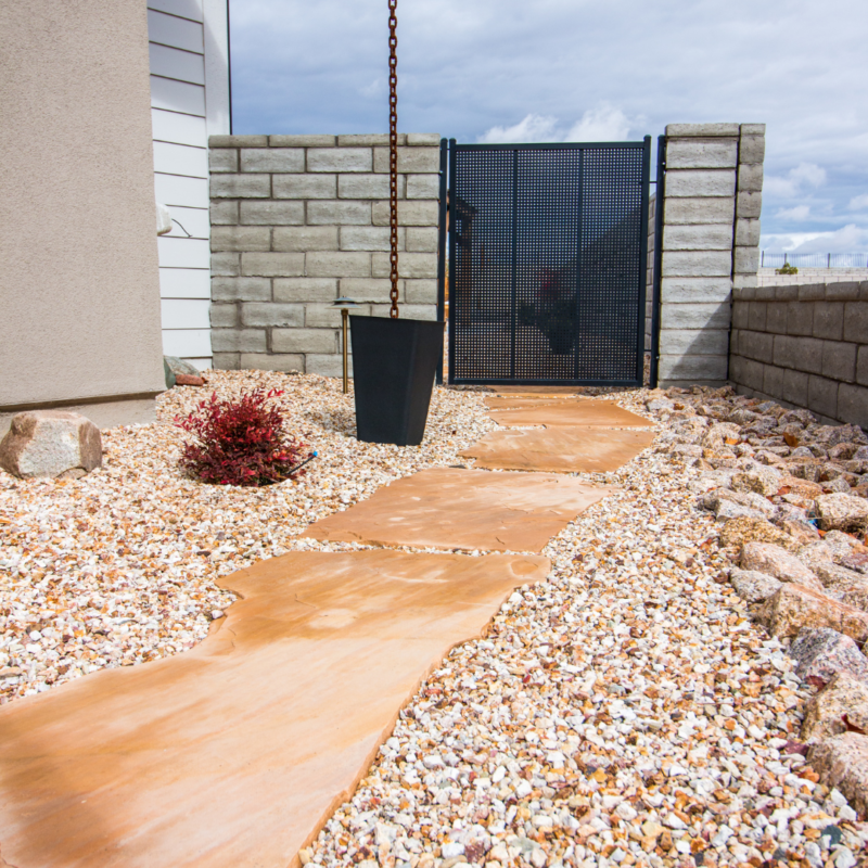 A landscaped outdoor area at the Ray Residence features a stone pathway leading to a black metal gate. The ground is covered in small colorful pebbles, and next to the path, a small red shrub adds vibrancy. A gray block wall partially encloses the serene space.