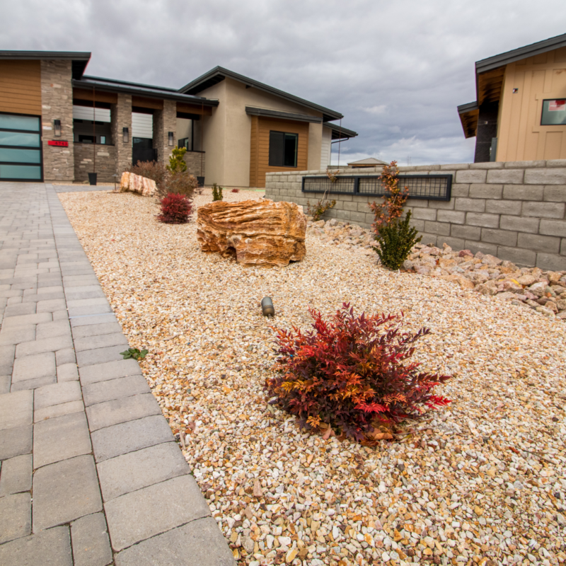 Ray Residence: Modern house with a stone and gravel front yard, featuring various bushes and large rocks. The property includes a stone driveway and a retaining wall on the right. Sky is overcast.