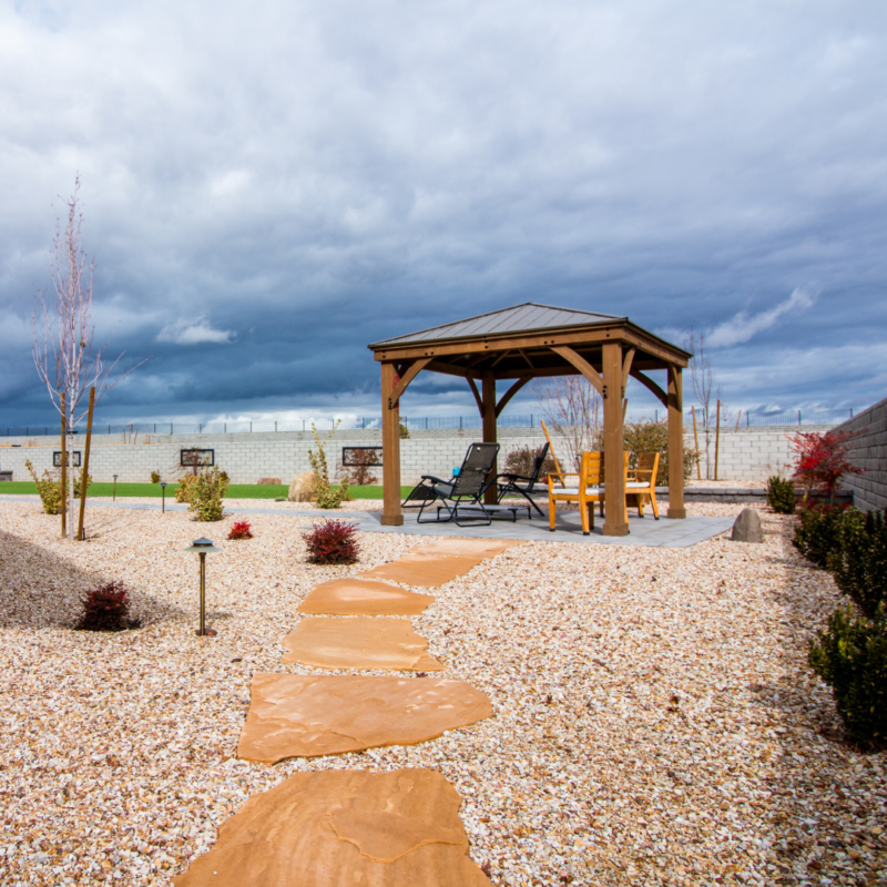 A backyard scene at the Ray Residence features a wooden gazebo on a stone patio, with lounge chairs and a small table. A stone path leads to the gazebo, bordered by decorative gravel and small plants. The sky is cloudy, suggesting an impending storm.