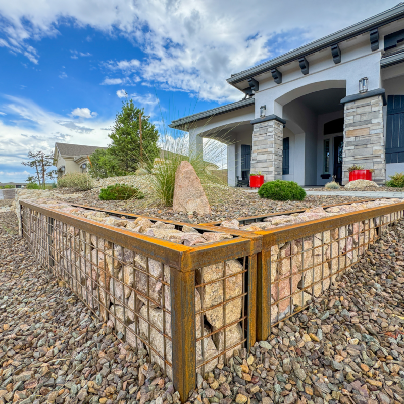 The Hutchinson Residence&#039;s modern landscaped garden features a raised bed made of rust-colored metal and wire mesh filled with rocks, surrounded by pebble ground cover. In the background, a house with arched doorways, stone accents, and red planters is visible under a blue sky with clouds.