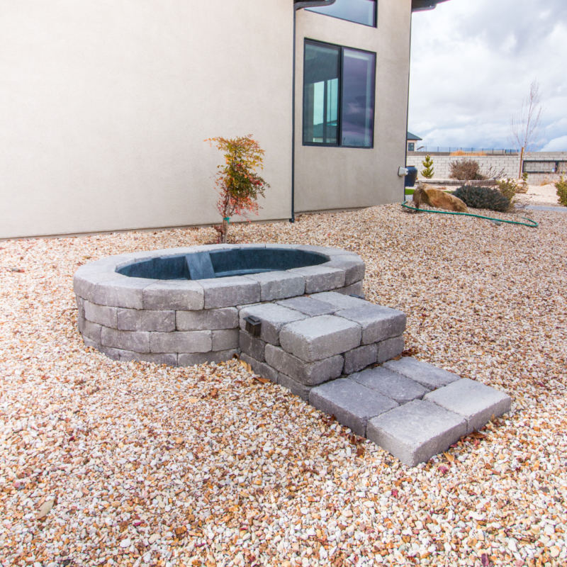 A small circular stone hot tub is built into a gravel-filled yard at the picturesque Ray Residence. Stone steps lead up to the hot tub. The background features a contemporary house with beige walls and large windows. A few small plants and a hose are visible in the yard.