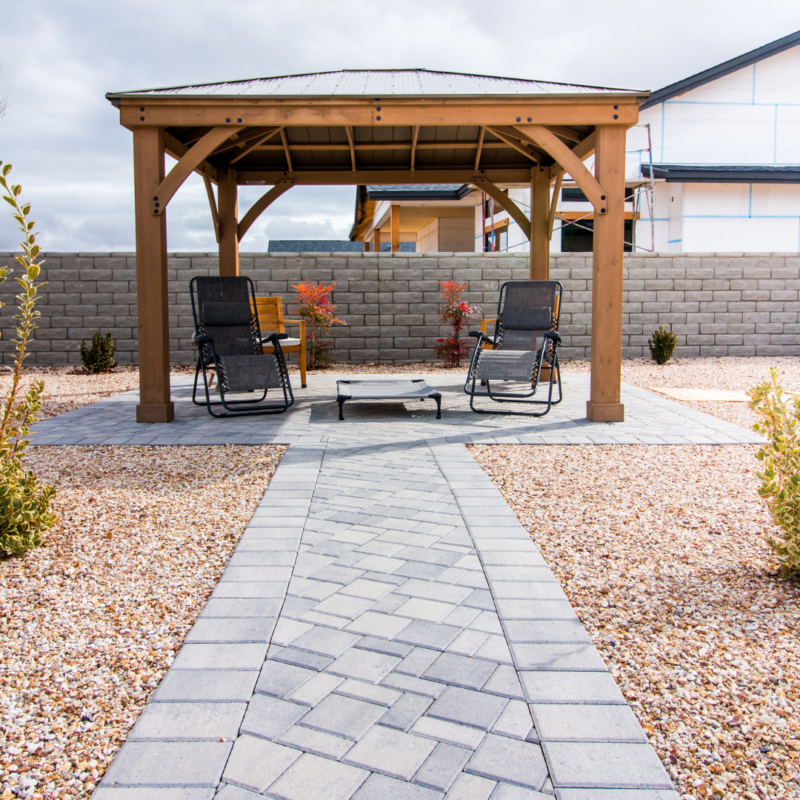 The Ray Residence features a backyard patio with a wooden gazebo. Inside the gazebo are two reclining chairs facing a fire pit on a paved surface. The surroundings are landscaped with gravel, small shrubs, and a stone pathway leading to the gazebo. A stone wall surrounds the yard.