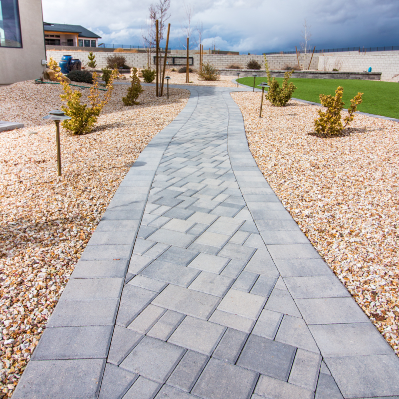 A winding stone pathway made of gray pavers, bordered by small rocks and shrubs, leads through a manicured garden at Ray Residence, with a green lawn on the right. In the background, there&#039;s a building and a high fence. The sky above is cloudy.