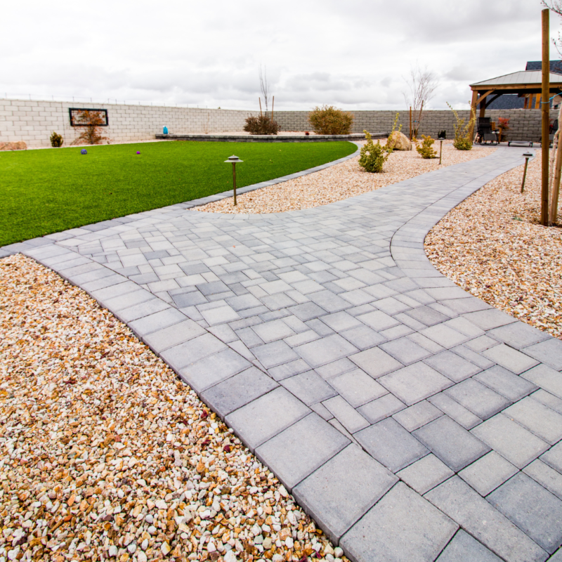 The Ray Residence backyard features a gray stone pathway winding through a gravel landscape with minimal vegetation. On one side of the path is a well-maintained green lawn, and on the other, there&#039;s a rock garden with a pergola in the background. The sky is overcast.