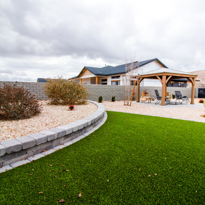 A modern backyard at the Ray Residence features a well-maintained lawn, a stone retaining wall, and a garden bed with shrubs. There&#039;s a wooden gazebo with outdoor seating and a table. The sky is overcast, but the scene feels tidy and inviting.