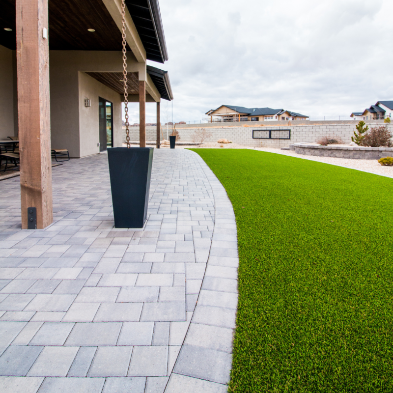 A modern backyard at the Ray Residence features a covered patio with a chain downspout, large rectangular planters, and a paved walkway. Adjacent to the walkway is a neatly manicured lawn, bordered by a stone wall and desert landscaping in the background under a cloudy sky.