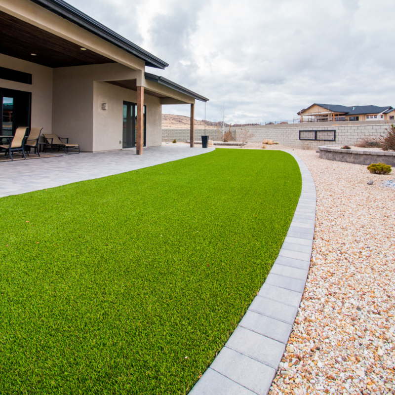 The Ray Residence boasts a backyard featuring a covered patio with lounge chairs and a neatly maintained artificial grass lawn. The lawn is bordered by a stone pathway and a gravel landscape with a retaining wall, with houses visible in the background under an overcast sky.