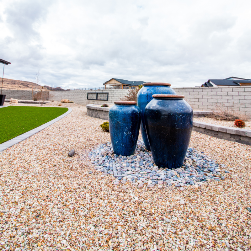 At the Ray Residence, three large, dark blue ceramic pots of varying heights are arranged in the middle of a pebble-covered landscape. They rest on a bed of small, multicolored rocks, with a well-manicured green lawn and a paved pathway in the background.