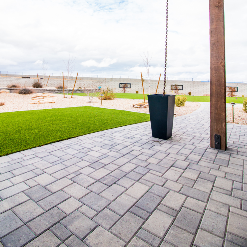 The Ray Residence backyard boasts a neatly paved patio, a hanging planter, and a well-maintained lawn area. The yard features a mix of artificial turf and desert landscaping with gravel, small plants, and a cinder block wall in the background.