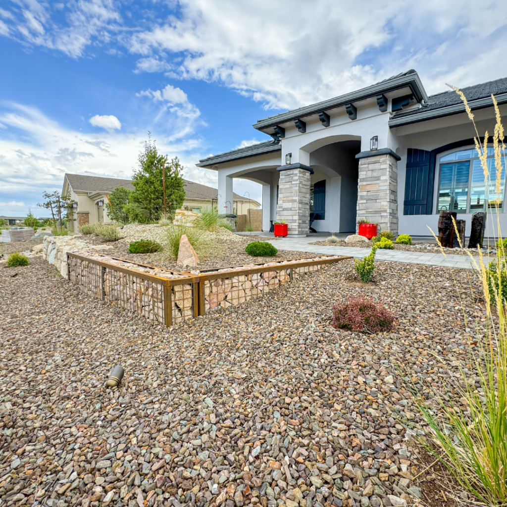 A modern house with a stone facade and wide porch is surrounded by a landscaped yard featuring decorative rocks, shrubs, and a small raised gabion rockwall garden bed. The sky is partly cloudy, adding a bright backdrop.