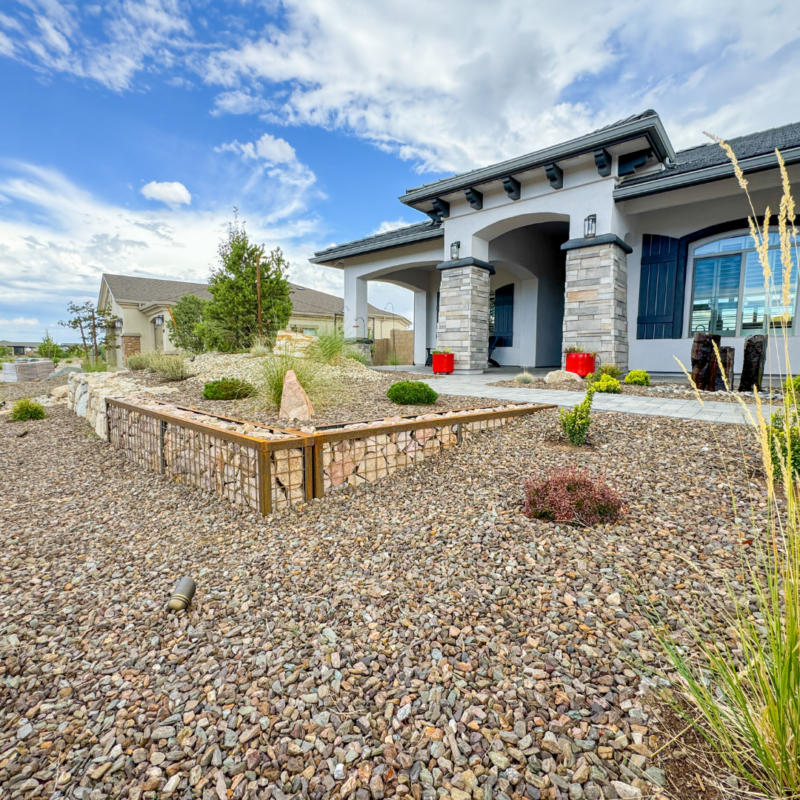 The Hutchinson Residence is a modern house with arched doorways, stone accents, and a covered porch, surrounded by a low-maintenance, gravel-filled yard with sparse vegetation and a few bushes. The sky is partly cloudy, creating a bright, pleasant atmosphere.