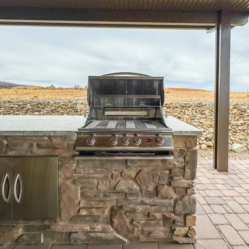 An outdoor built-in brick grill with a stainless steel barbecue setup and auto draft feature. The grill area is under a covered patio, with a stone countertop and cabinet space beneath. The background shows a rocky terrain with an open field and a cloudy sky.