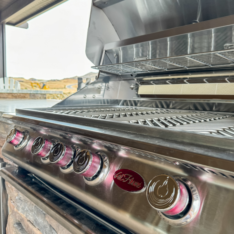 Close-up of a shiny, stainless steel Koehler outdoor grill with four burners, each boasting a pink-lit control knob. The grill lid is open, revealing the grilling area. In the background, part of a residence and landscape can be seen.