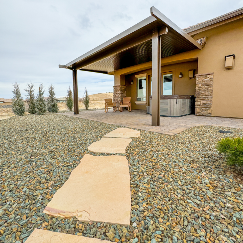 The Koehler Residence is a modern house with a covered patio, outdoor seating, and a hot tub. The yard features a pebble ground cover with stepping stones leading to the patio. Leafless trees are planted along the side of the house, and the sky is mostly cloudy.