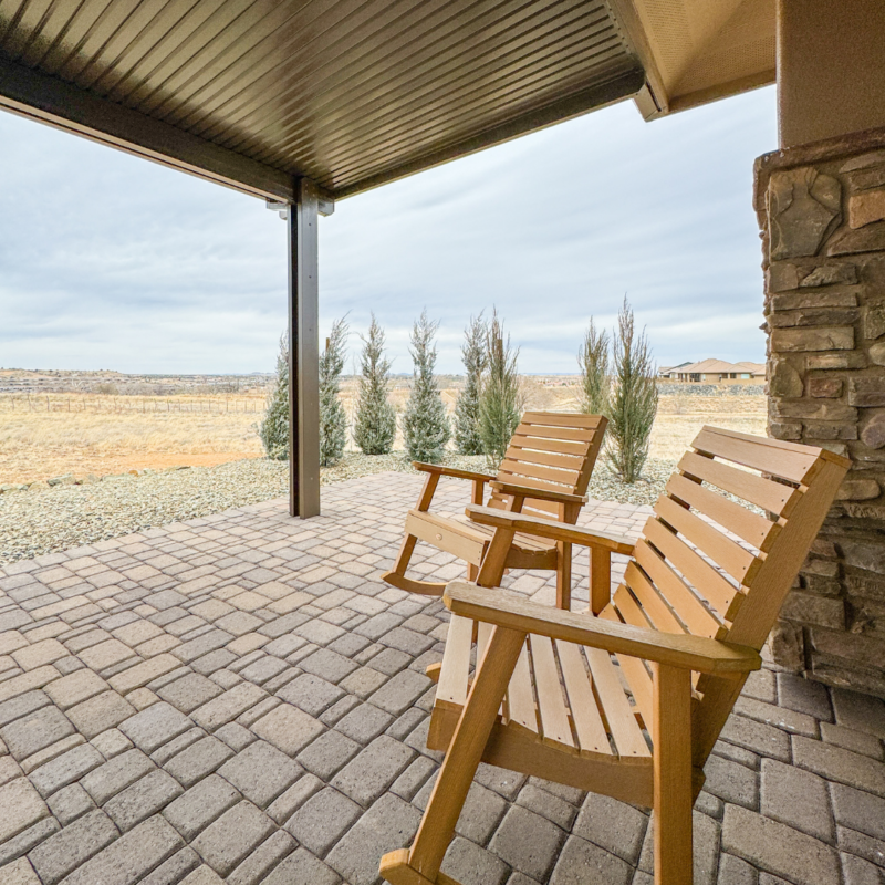 Two wooden chairs on a brick patio under a covered roof overlook the serene landscape of the Koehler Residence, with dry grass and shrubs in the distance. The patio is bordered by stone siding and offers a clear view of the open sky and distant homes.